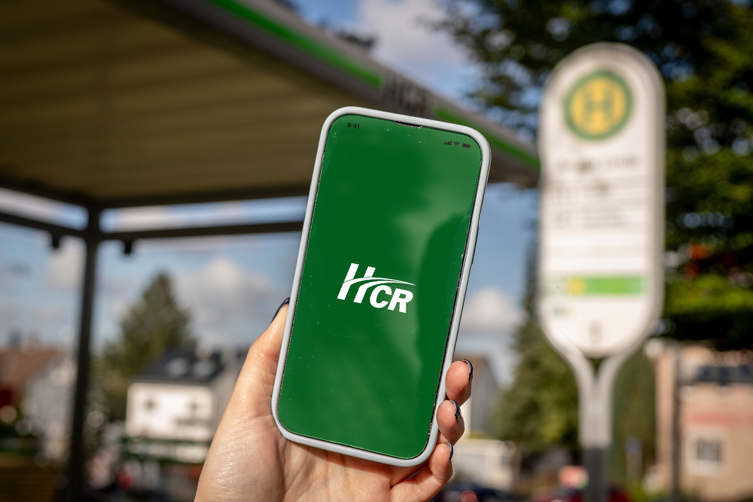 Eine Hand hält ein Smartphone mit der startenden HCR App. Unscharf im Hintergrund ist eine Haltstelle zu sehen.