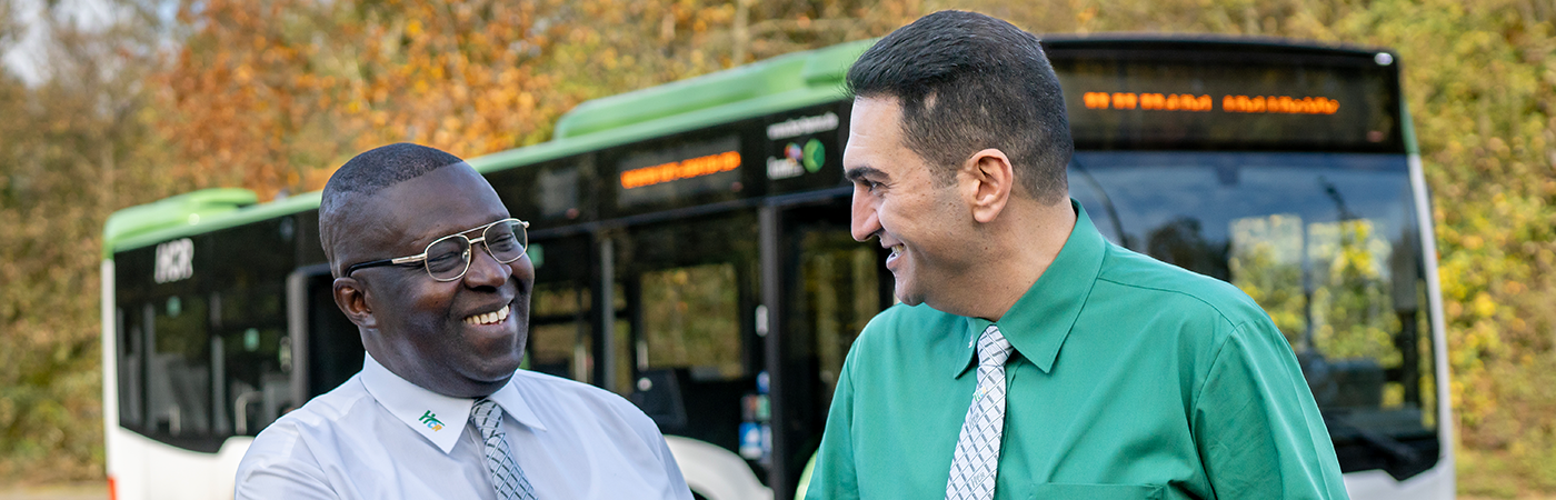 Kampagnenmotiv der HCR-Personalkampagne. Zwei Busfahrer schauen sich lächelnd an. Im Hintergrund ist ein HCR-Bus zu sehen.