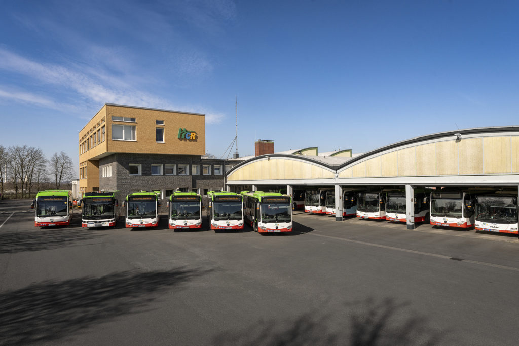 HCR-Betriebshof mit sechs Bussen, die vor dem Verwaltungsgebäude stehen. Weitere Busfronten sind in den Fahrzeughallen zu sehen.