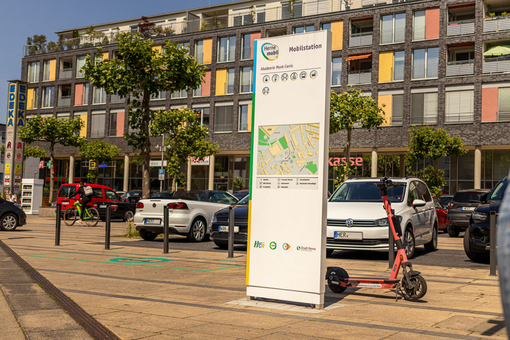 Stele der Mobilstation an der Akademie Mont-Cenis. Neben der Stele ist ein E-Scooter abgestellt.