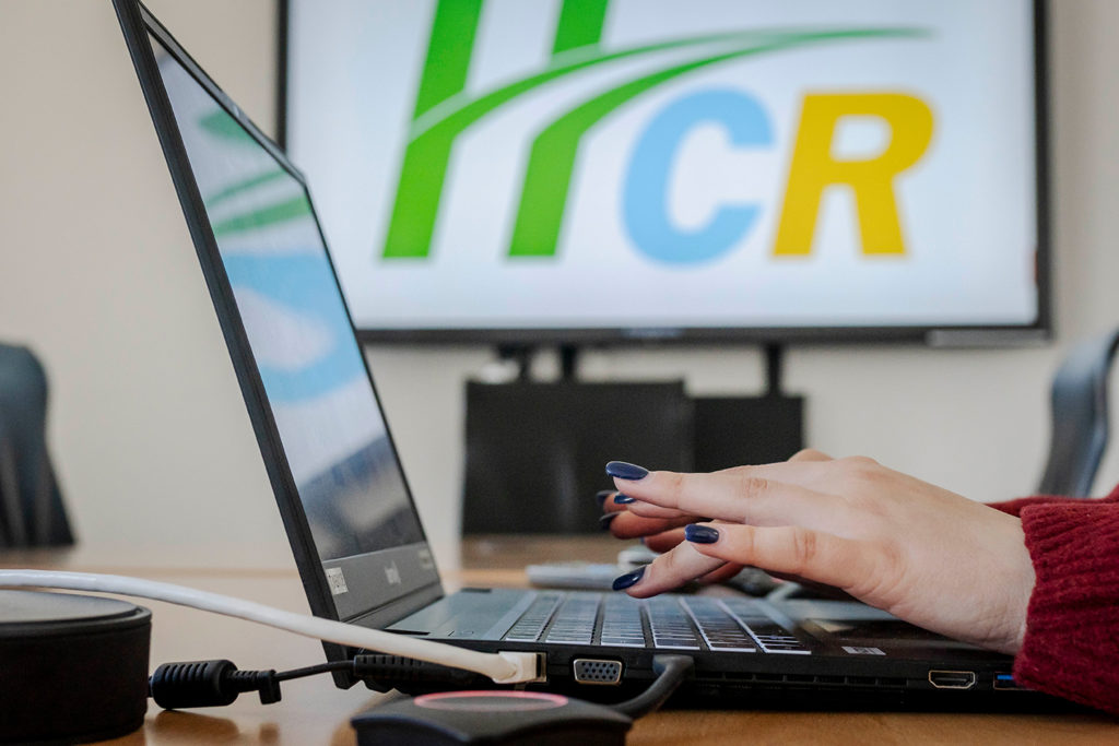 Zwei Hände tippen auf einem Notebook. Im Hintergrund ist ein großer Bildschirm mit dem HCR-Logo zu sehen.
