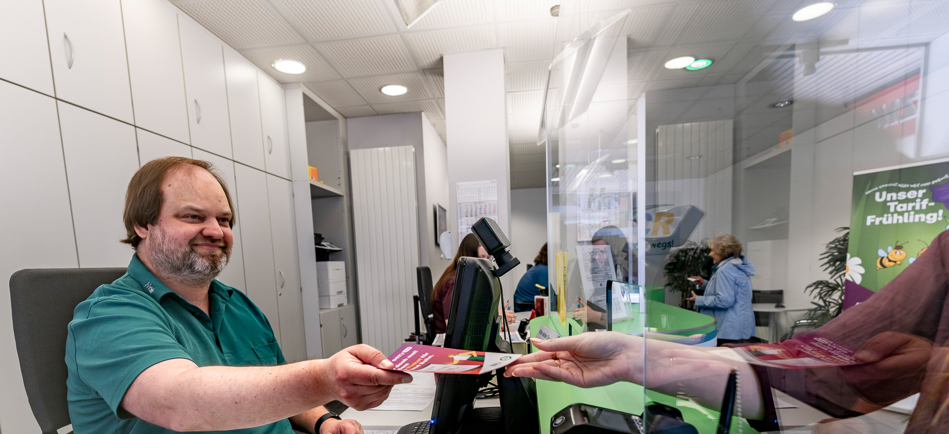 Im KundenCenter Herne-Mitte übergibt ein Mitarbeiter einer Kundin eine Broschüre, Im Hintergrund sind weitere Mitarbeiter und eine Kundin zu sehen.